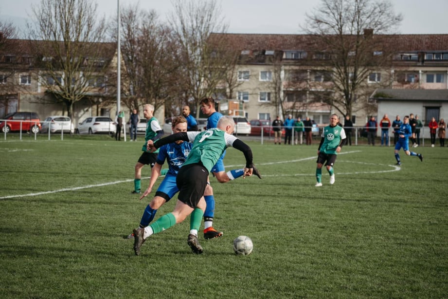 Fußballspieler im Zweikampf, im Hintergrund noch mehr Spieler und Zuschauer
